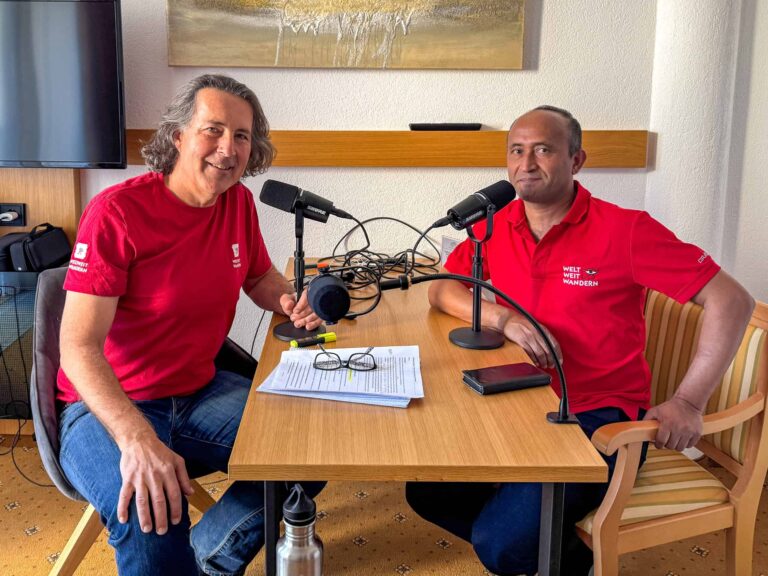Auf dem Bild sitzen zwei Männer an einem Tisch, die in ein Mikrofon sprechen, als würden sie ein Interview oder einen Podcast aufnehmen. Beide tragen rote T-Shirts mit dem Aufdruck „WELT WEIT WANDERN“. Auf dem Tisch liegen einige Papiere, eine Brille, ein Marker und ein Smartphone. Im Hintergrund ist eine helle Wand mit einem Bild sowie ein Fernseher an der Wand zu sehen. Das Setting wirkt freundlich und professionell.