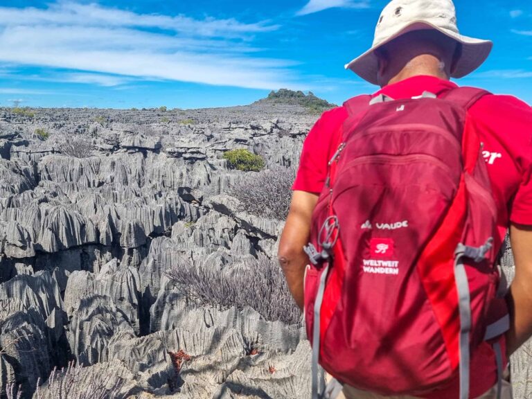 Ein Mann mit rotem T-Shirt, beigem Sonnenhut und rotem Rucksack steht am rechten Bildrand und blickt auf eine raue, zerklüftete Felslandschaft, die sich weit nach hinten erstreckt. Über ihm ist ein blauer Himmel mit wenigen Wolken zu sehen. Die Szene wirkt sonnig und klar.